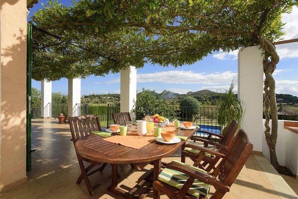 Charming covered terrace with a wooden outdoor dining table, surrounded by lush greenery and open views of the countryside and mountains. A perfect space to enjoy al fresco breakfasts in a peaceful and private setting, with a pool and garden enhancing the Mediterranean experience.