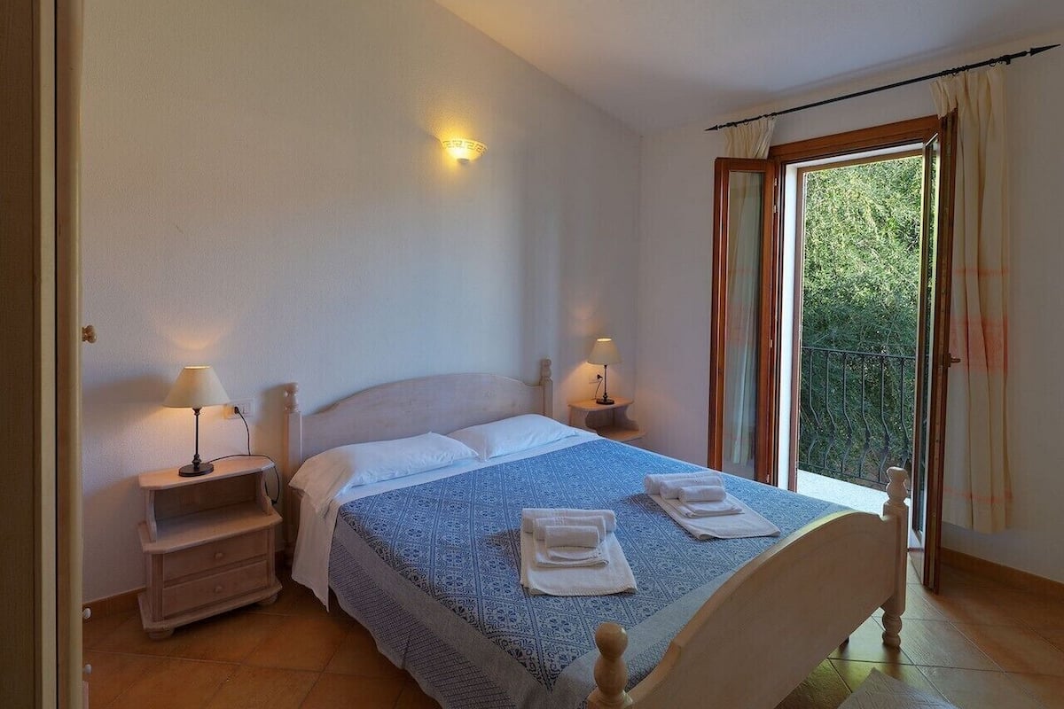 Quarto com cama, duas mesas de cabeceira com abajures, janela com cortinas e vista para o verde lá fora.