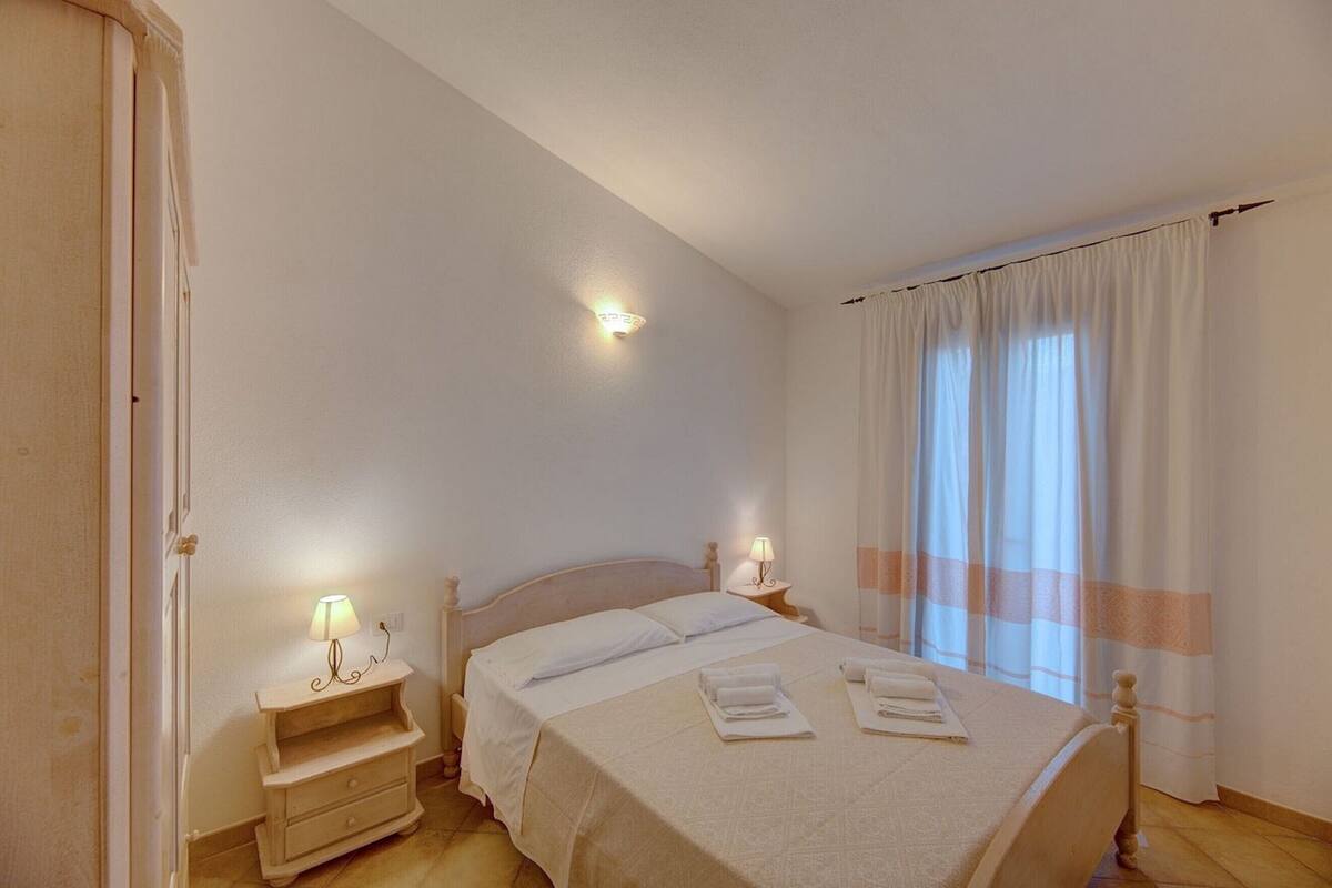 Quarto com cama, abajures, mesa de cabeceira e janela com cortinas.