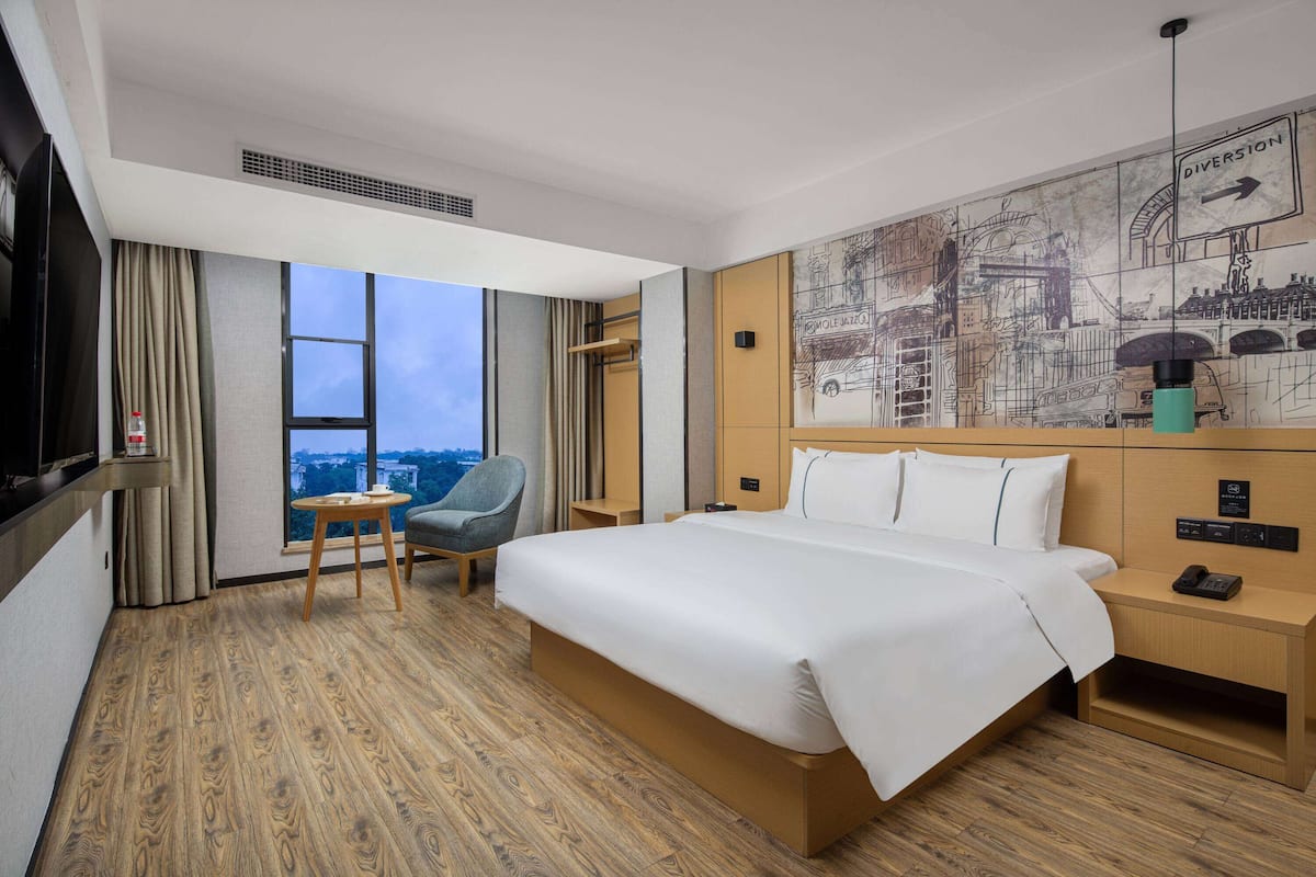 A hotel room with a large bed, a small table, a chair, a TV, a window with curtains, and a wall with a cityscape mural.