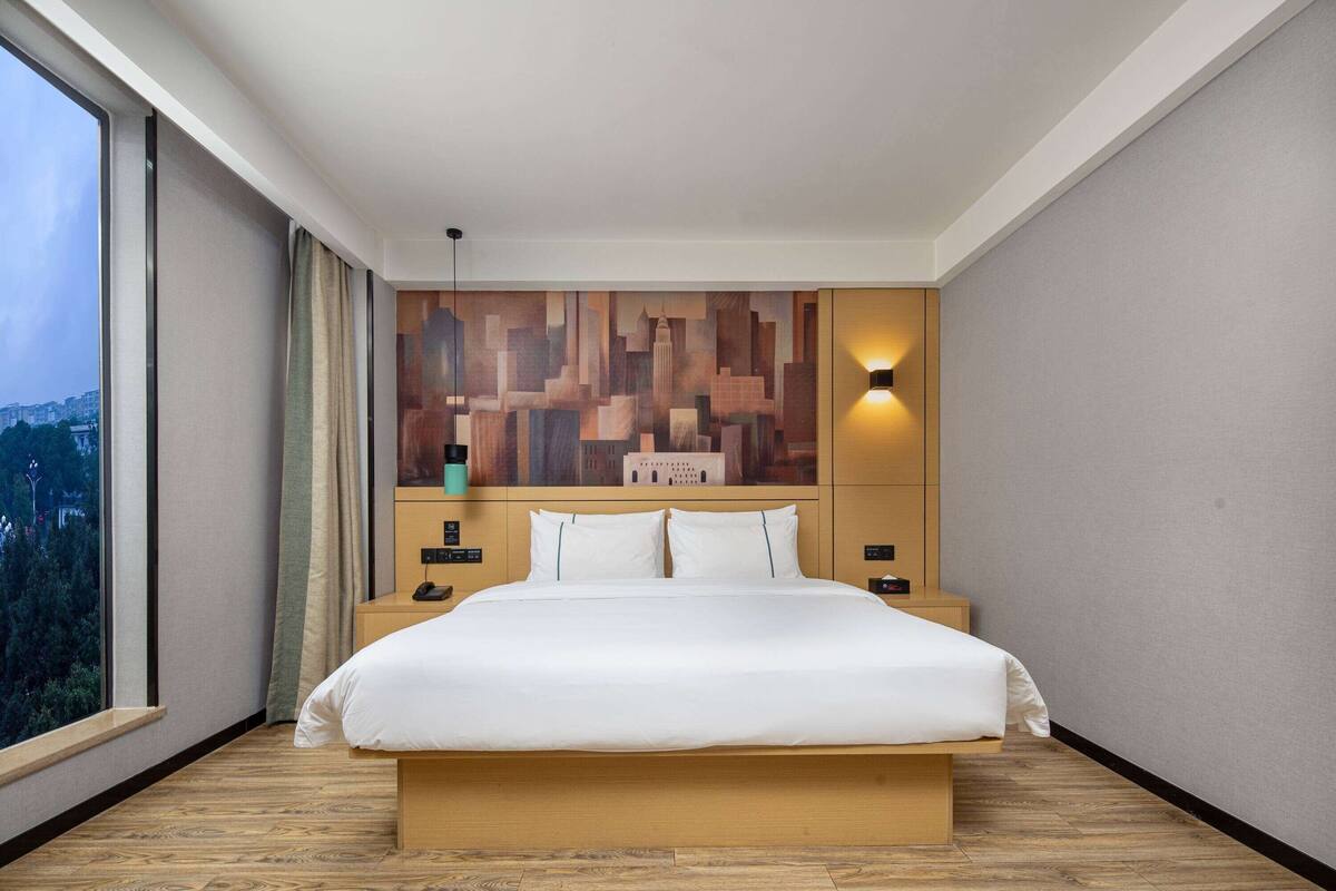A modern hotel room with a large bed, a cityscape view, a wall-mounted artwork, and a bedside lamp.