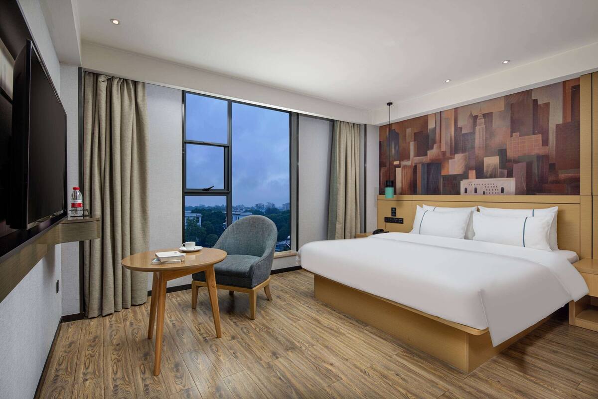 A hotel room with a large bed, a chair, a small table, a TV, and a cityscape view through the window.
