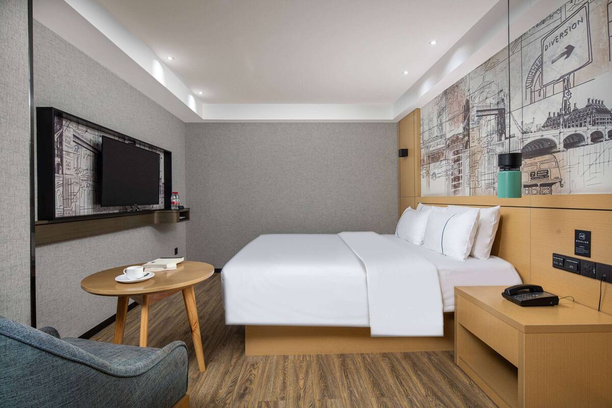 A modern hotel room with a bed, a small table, a chair, a TV, and a wall art piece.