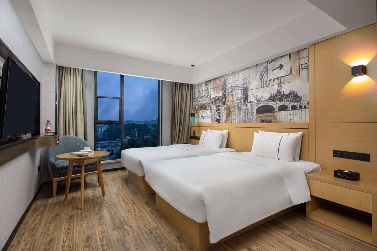 A hotel room with two beds, a small table, a chair, a TV, and a large window with a city view.