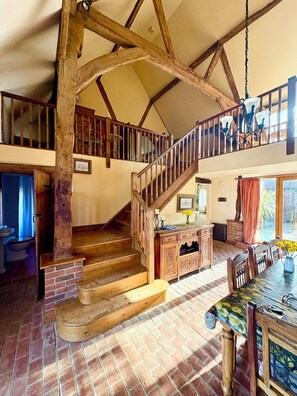 A spacious loft with exposed wooden beams, a wooden staircase, a dining area with a table and chairs, and a view of the outdoors.