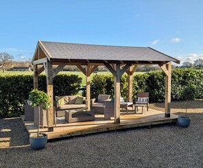 A wooden gazebo with a sloped roof, furnished with wicker sofas and a coffee table, surrounded by gravel and greenery.