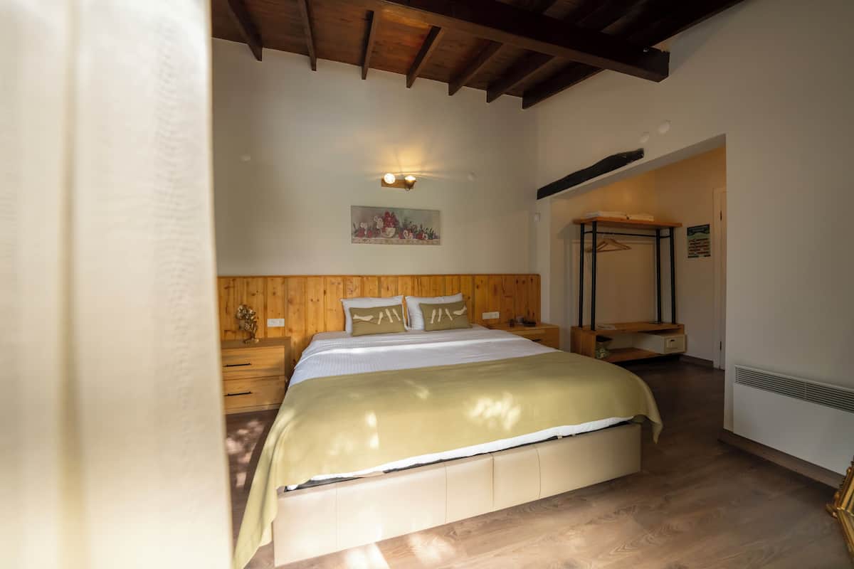 Un dormitorio con cabecera de madera, cama con colcha beige y blanca, mesita de noche, lámpara de pared y perchero de madera.
