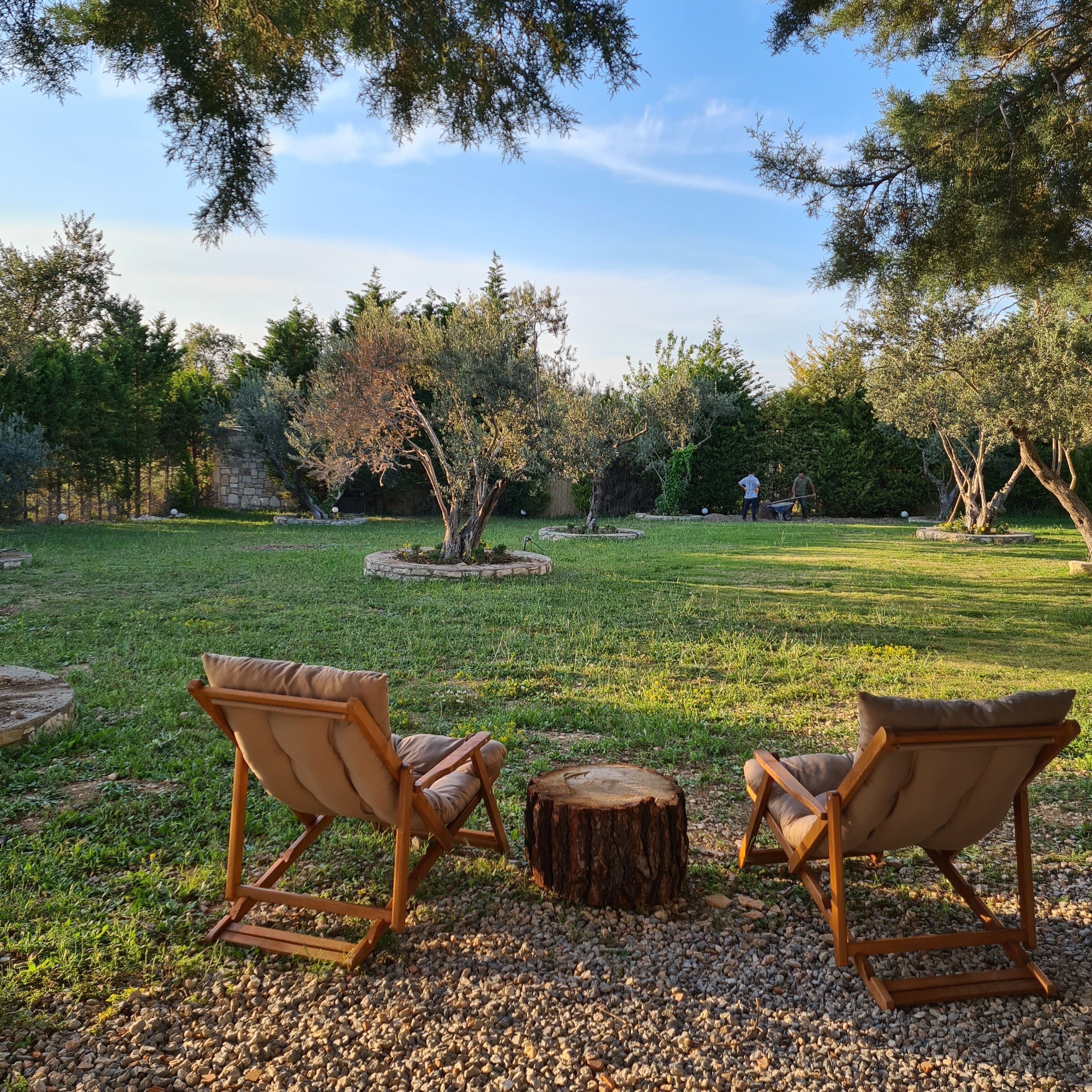 Two wooden lounge chairs facing a circular stone table in a grassy outdoor area with trees and a stone wall in the background.