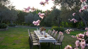 An outdoor dining area with a long table set for a meal, surrounded by chairs, in a garden with trees and flowering plants. - Bir dem Urla (Urla)