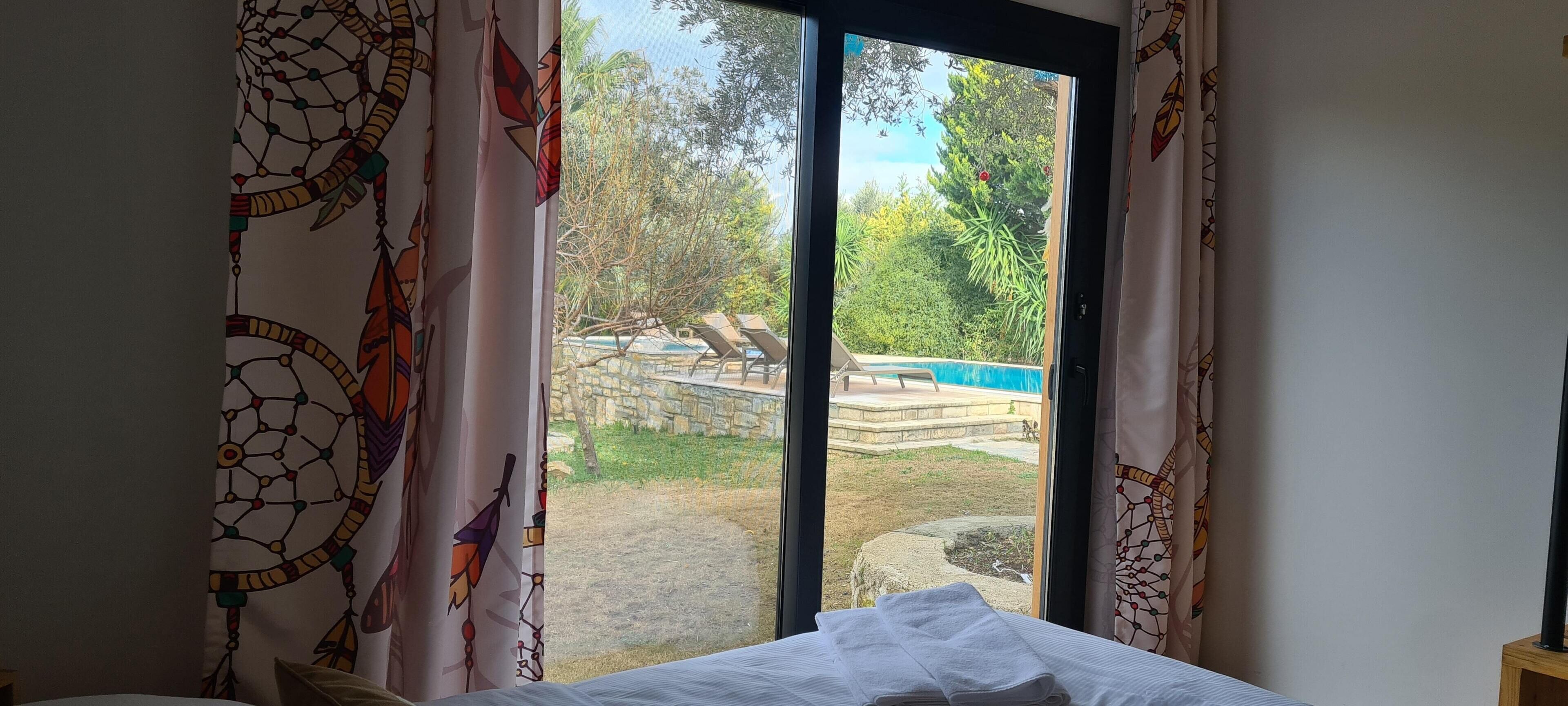 Una habitación con cama, ventana con cortinas y vistas a una piscina y jardín.