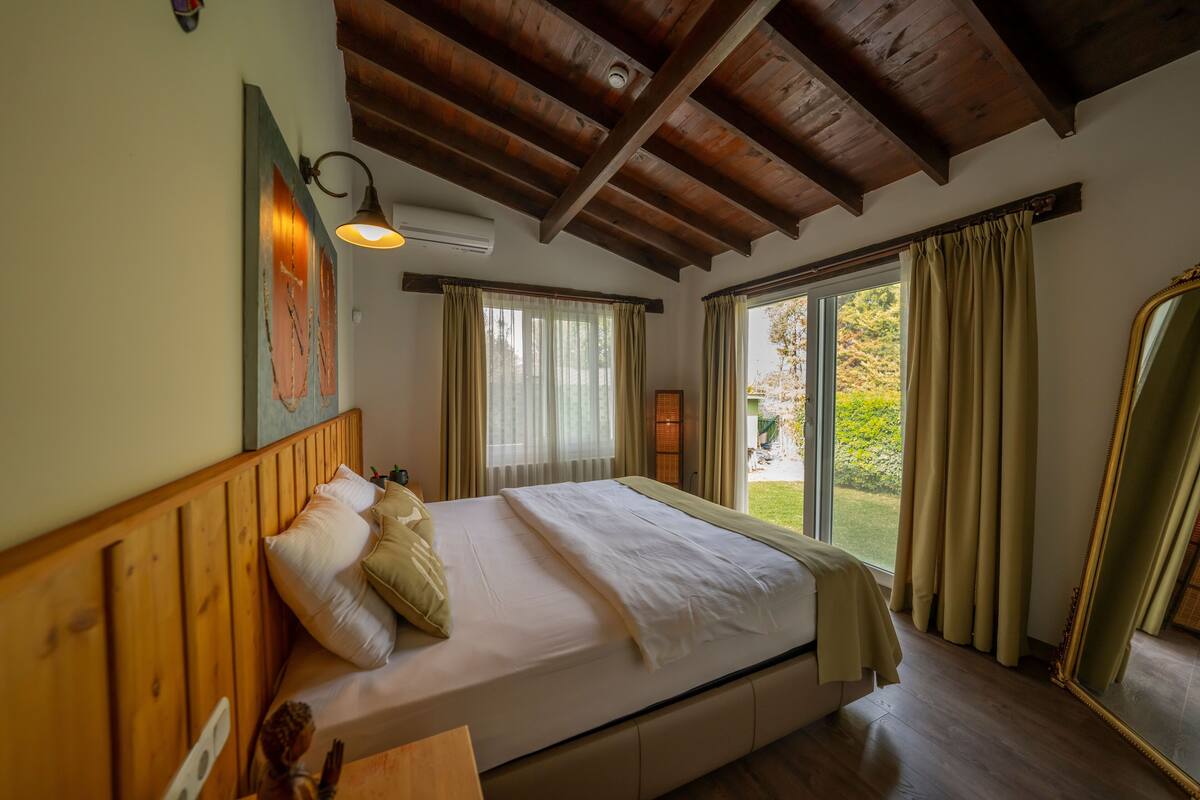 Un dormitorio con techo de madera, cama con ropa blanca, espejo grande y vista a zonas verdes.