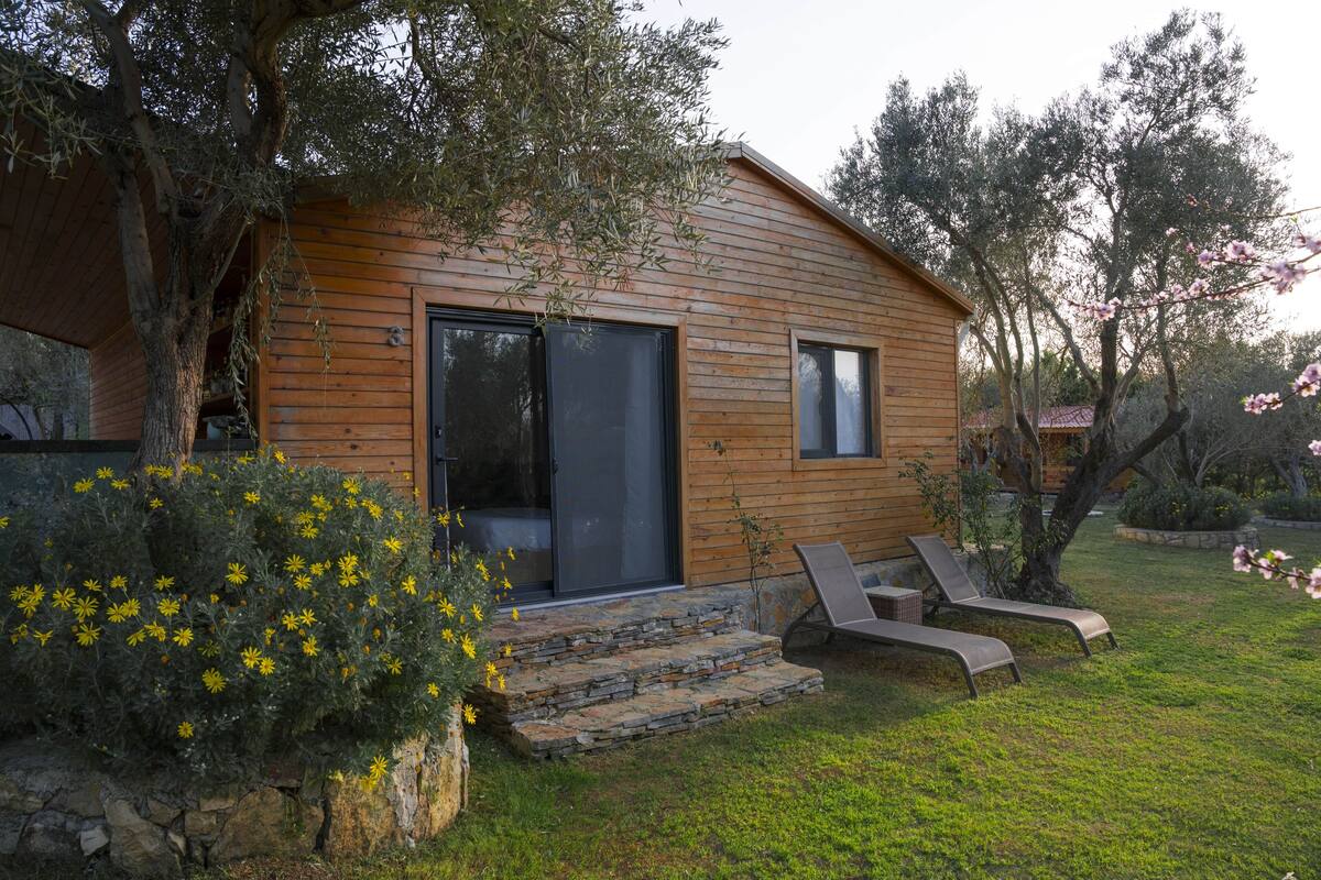 Una cabaña de madera con una escalera de piedra que lleva a una puerta, dos sillones y un jardín con flores amarillas.