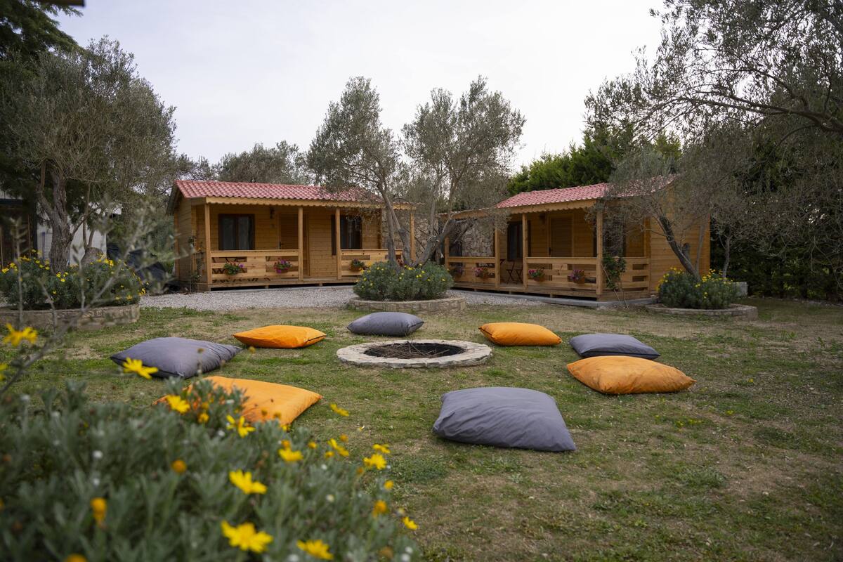 Una cabaña de madera rústica con techo de tejas rojas, rodeada por un jardín con diversas plantas y flores.
