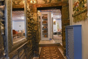Un pasillo con paredes de piedra, un tapete con diseño, un espejo y una puerta que da a una habitación con un aparador azul y una estantería con adornos.