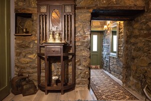 Un pasillo con paredes de piedra, un mueble de madera con cristalería, una alfombra estampada y una puerta verde al final.