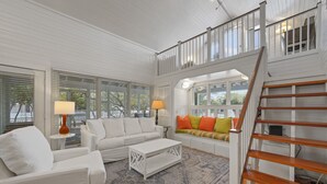 A spacious living room with a white sofa, a coffee table, and a colorful bench. A wooden staircase leads to an upper level. - Directly Across From Beach With Gulf Views in Seaside, FL + 2 Adult Bikes! (Santa Rosa Beach)