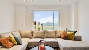 A modern living room with a beige sectional sofa, a round coffee table, and a large window offering a view of a green field. - Vacation home Staedenbergh in rural Staden! 10 pers (Staden)
