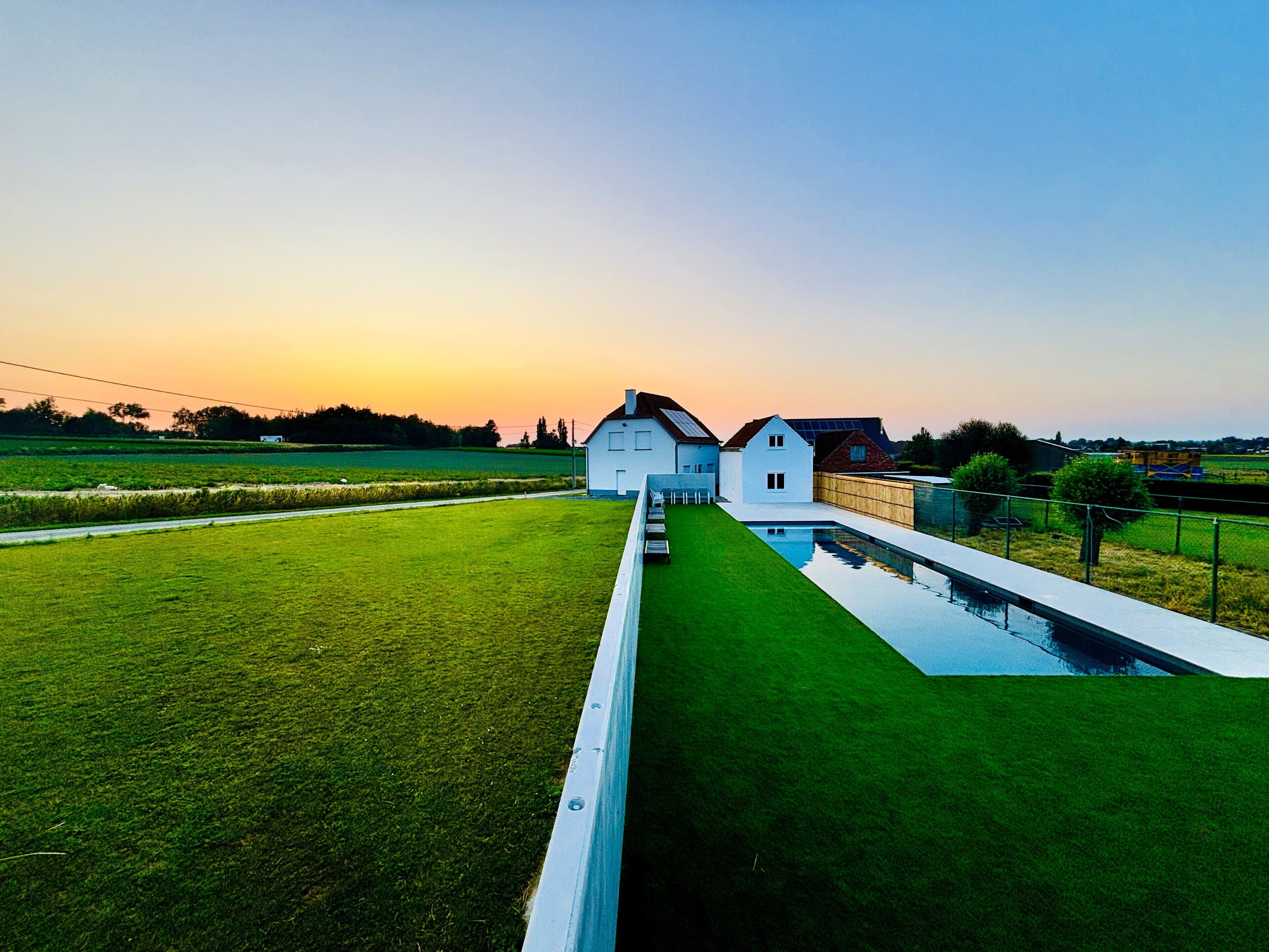A modern house with a swimming pool, surrounded by green fields and a clear sky.