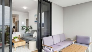 En modern lägenhet med ett vardagsrum som har en grå soffa, ett soffbord i trä och en lila fåtölj. I bakgrunden syns ett kök, och ett stort fönster erbjuder utsikt över staden.