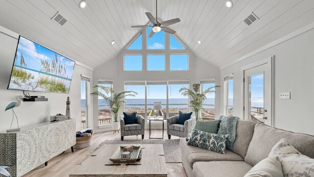 A spacious living room with a high ceiling, large windows offering a view of the beach, a flat-screen TV mounted on the wall, and a cozy seating area with a sofa and armchairs. - Ocean Dreams Beachfront Home Stunning Views Like New Sleeps 10 4 BR 3 Full Baths (Gulf Shores)