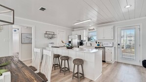 A modern kitchen with white cabinetry, a central island with stools, and a dining area with a wooden table and white chairs. - Ocean Dreams Beachfront Home Stunning Views Like New Sleeps 10 4 BR 3 Full Baths (Gulf Shores)