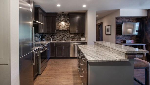 A modern kitchen with dark wood cabinets, stainless steel appliances, a marble countertop, and a stone backsplash. - Zephyr Mountain Lodge, Condo | 3 bedroom Ski Slope Views (Premium rated Condo 2407) (Winter Park)