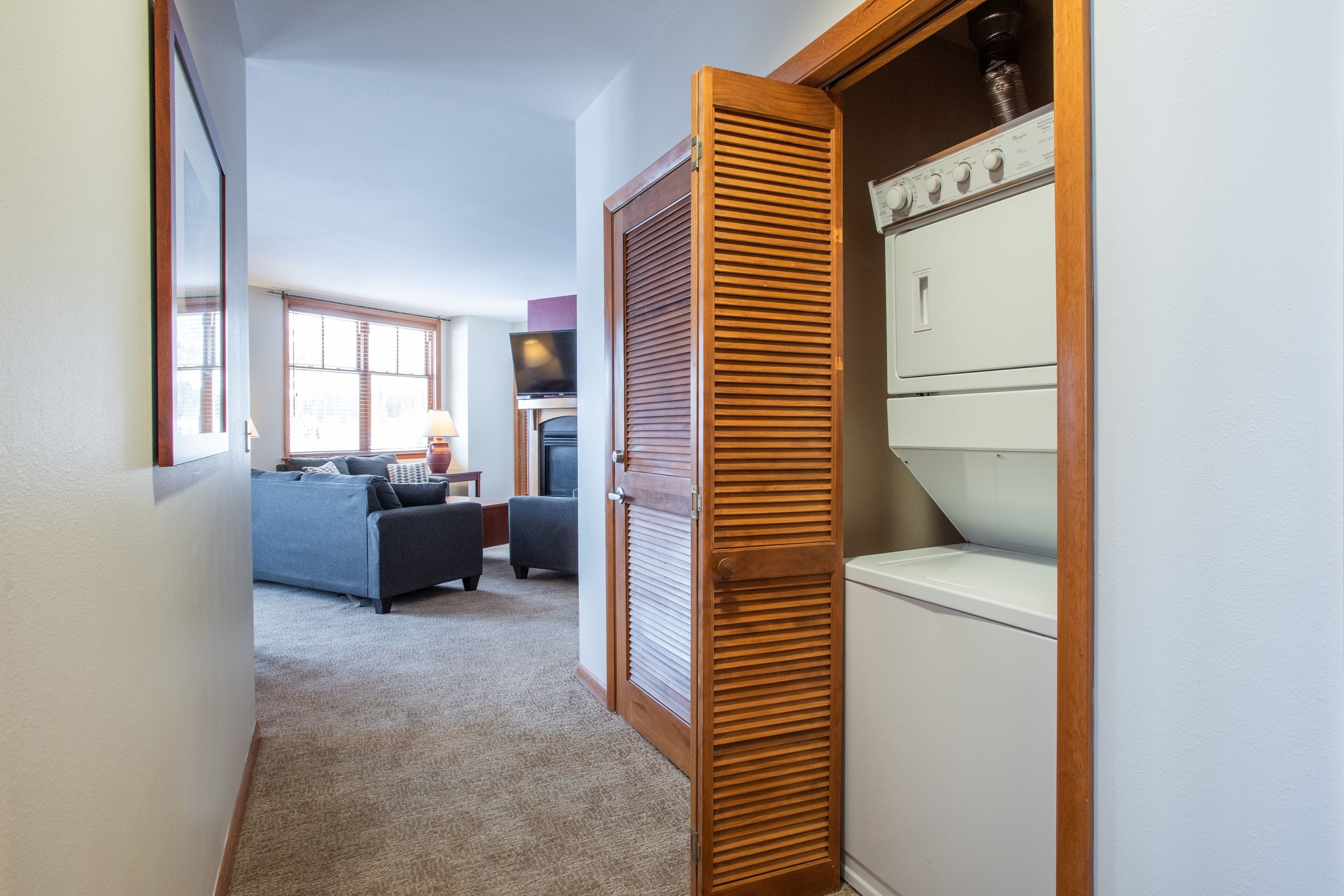 Un pasillo con una puerta de madera, una lavadora blanca y una sala de estar con sofá y televisor.