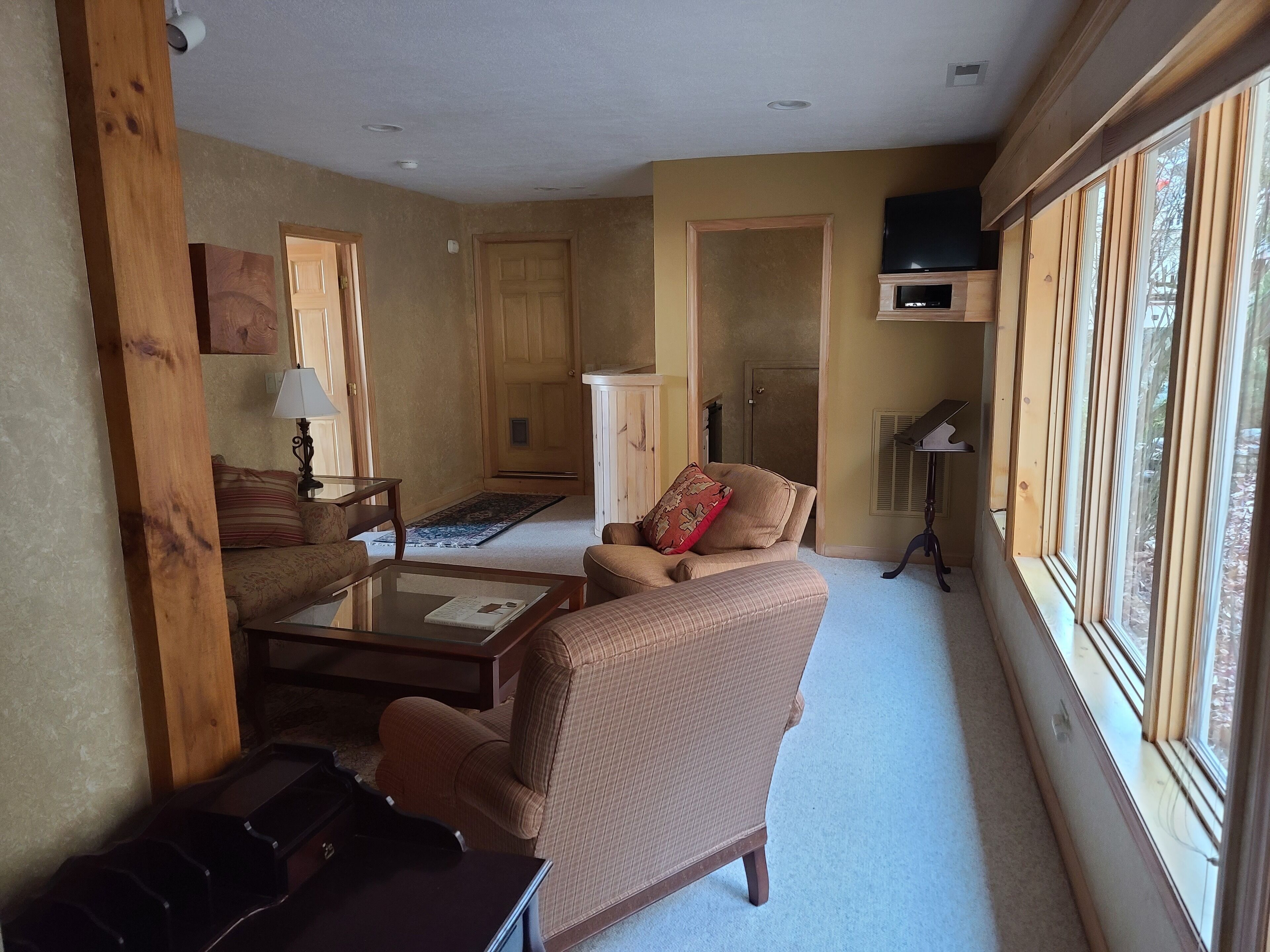 Un salon comprenant un canapé, un fauteuil et une table basse. On y trouve une fenêtre avec des stores, une lampe et un téléviseur posé sur un meuble.