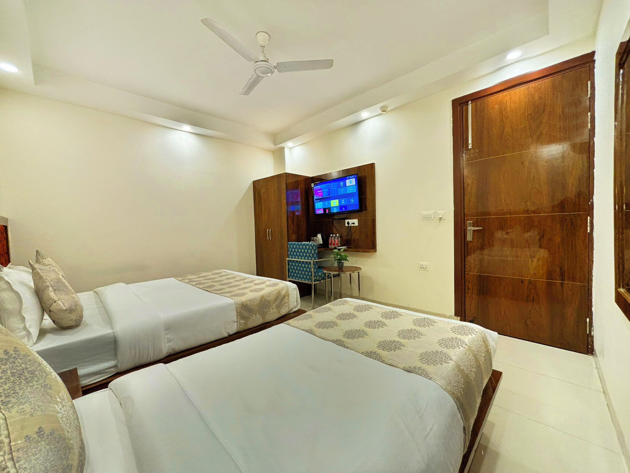 A hotel room with two beds, a TV mounted on the wall, a ceiling fan, and wooden wardrobes.