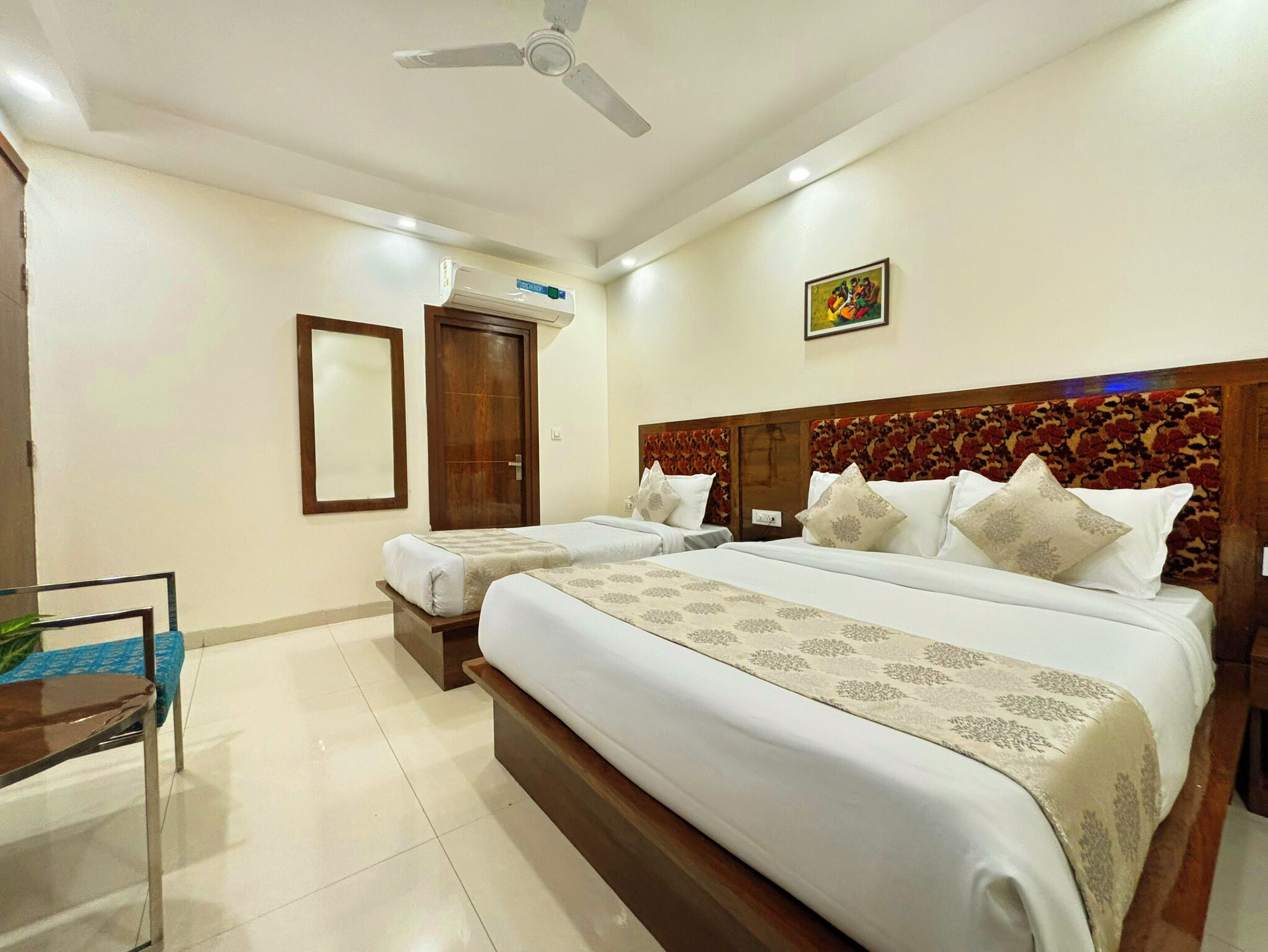 A hotel room with a large bed, two smaller beds, a ceiling fan, an air conditioning unit, a wooden headboard, and a framed picture on the wall.