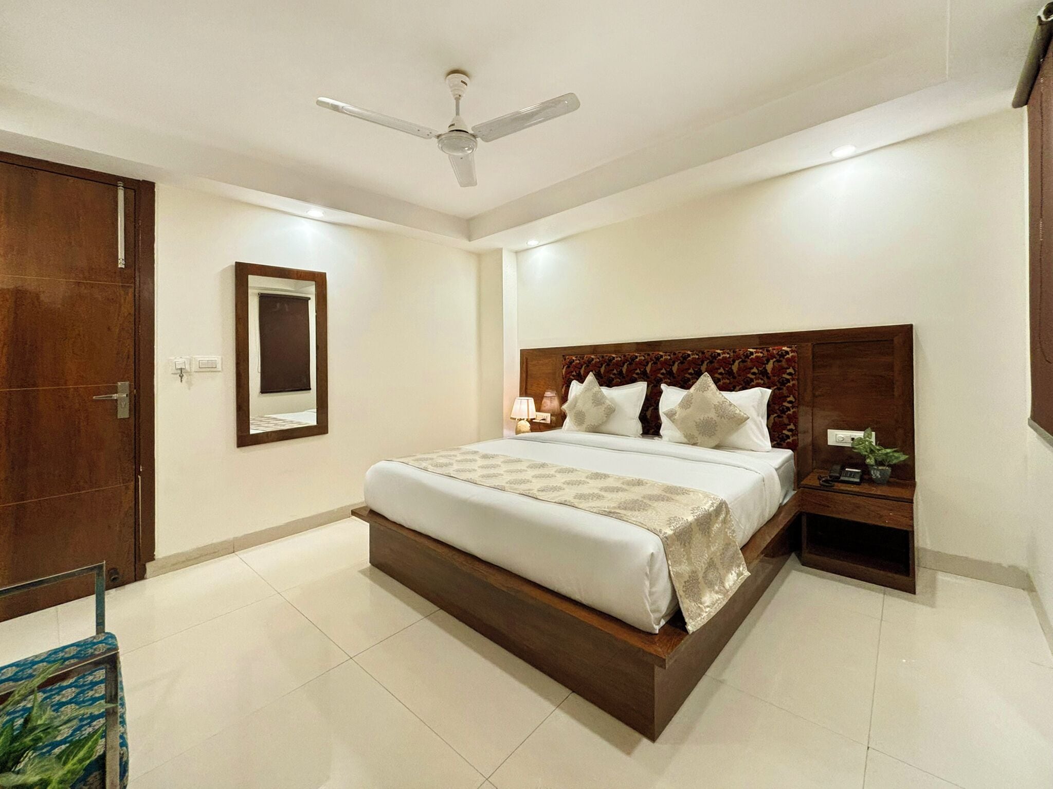 Quarto de hotel com uma cama grande, cabeceira de madeira, mesa de cabeceira, ventilador de teto e um espelho na parede.