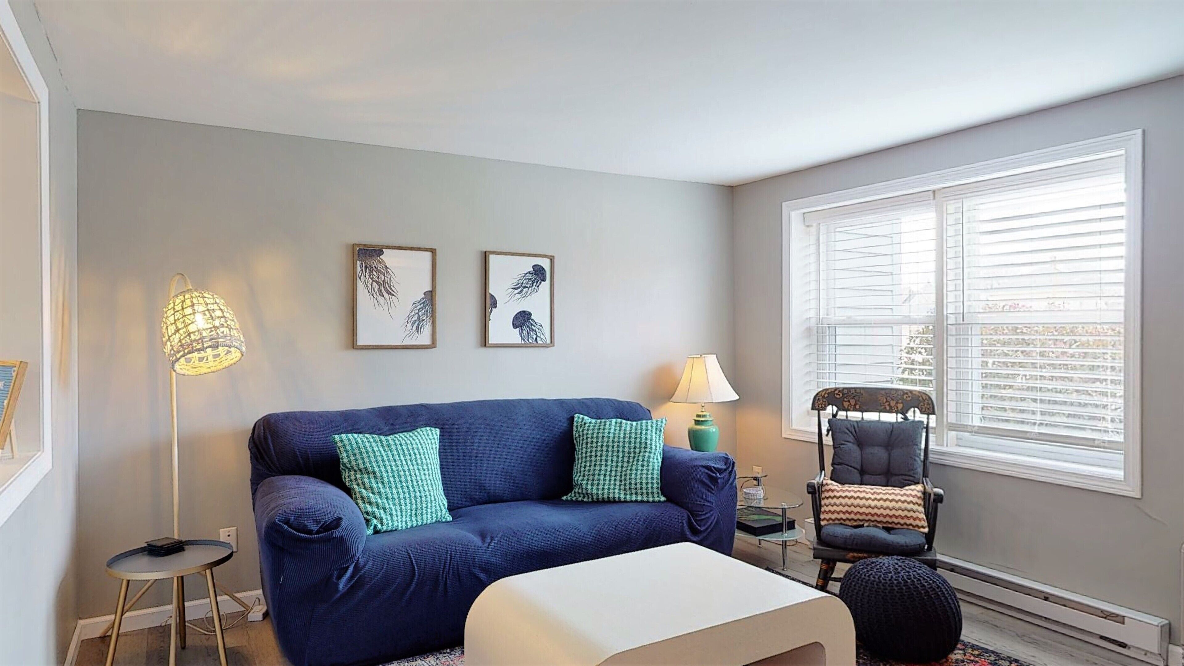 Un salon avec un canapé bleu, deux coussins verts, un pouf blanc, une chaise noire avec un coussin à motifs, une lampe sur pied, une lampe de table et deux tableaux encadrés accrochés au mur.