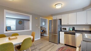 A modern kitchen with stainless steel appliances, a dining area with a white table and green chairs, and a view into another room with a wicker chair and a clock.