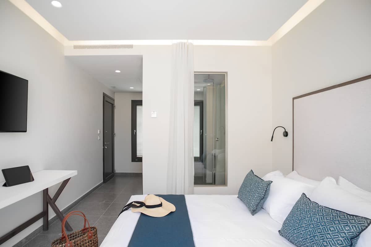 A modern hotel room with a bed, a desk, a TV, and a bathroom visible through a glass door.