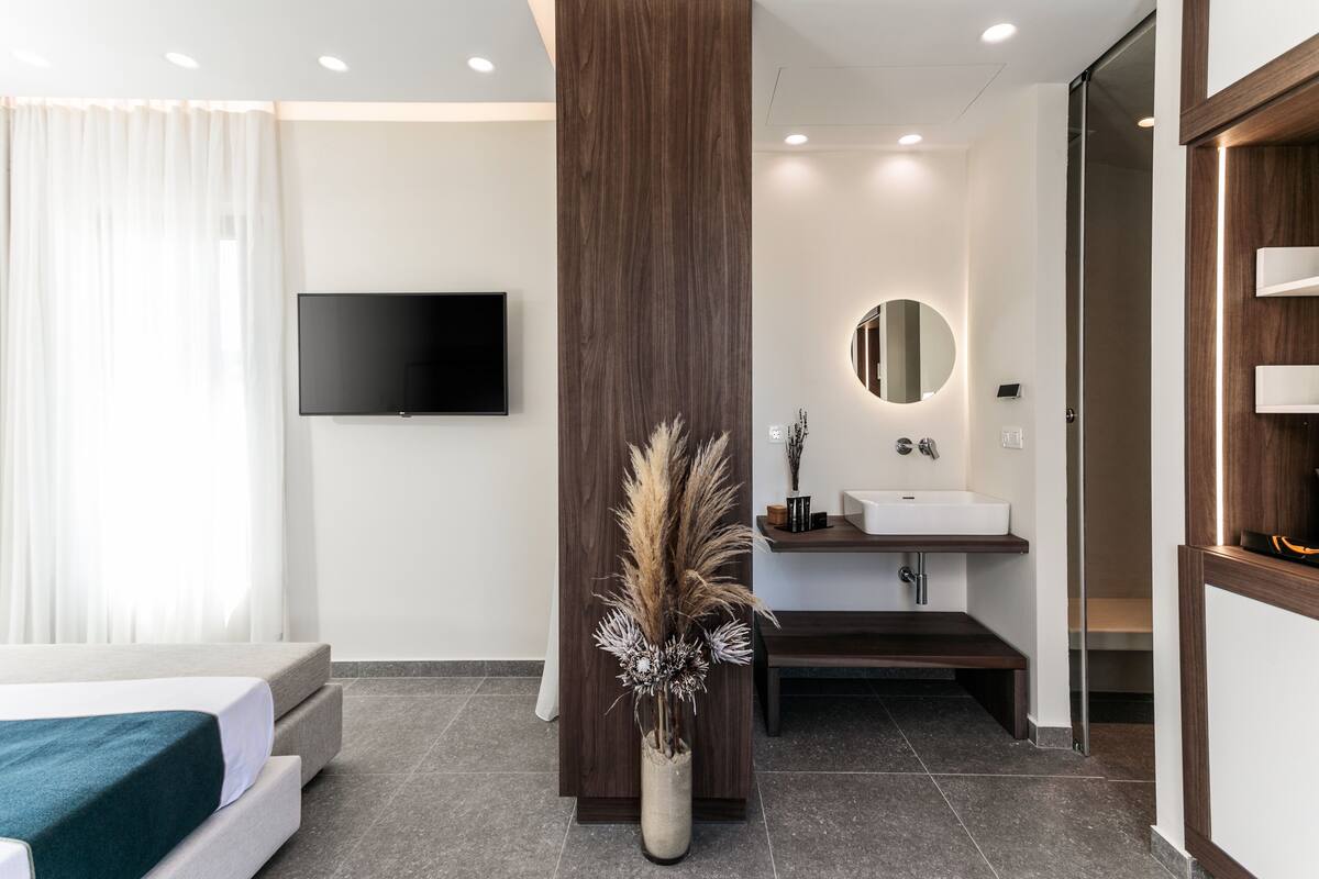 A modern hotel room with a bed, a flat-screen TV, a wooden column, a bathroom with a sink and mirror, and a vase with dried plants.