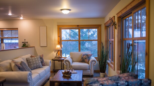 A cozy living room with a sofa, armchair, and a coffee table. There is a window with a view of trees and a lamp on a side table. - Beautiful 6-bed, 4.5-bath with private outdoor space.  5min walk to Main Street. (Lake Placid)