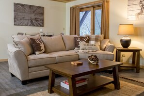 A living room with a beige sofa, a wooden coffee table, and a lamp on a side table. - Beautiful 6-bed, 4.5-bath with private outdoor space. 5min walk to Main Street. (Lake Placid)