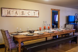 Uma sala de jantar com uma longa mesa de madeira posta para refeição, uma cadeira xadrez e um banco. Uma placa com o nome "MARCY" está pendurada na parede.