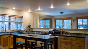 A modern kitchen with wooden cabinets, a large island with stools, and a view of the outdoors through large windows. - Beautiful 6-bed, 4.5-bath with private outdoor space.  5min walk to Main Street. (Lake Placid)