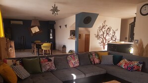 A modern living room with a grey sectional sofa, wooden coffee table, and decorative pillows. A dining area with a yellow chair and a wall-mounted clock is visible in the background.