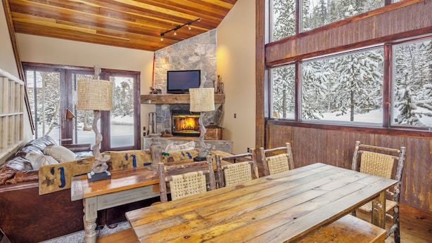 Interior acogedor de una cabaña con chimenea de piedra, muebles de madera y grandes ventanales con vistas a un bosque nevado.