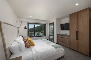 A modern bedroom with a large bed, a nightstand, a desk, a wardrobe, a TV, and a window with a view of trees.