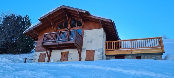 A chalet-style building with a wooden balcony and stone facade, surrounded by snow.