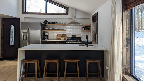 A modern kitchen with a large island, wooden stools, a stainless steel refrigerator, and a window with a view of a snowy landscape. - Acorn Bungalow & Lodge - Luxury Cabin, Catskills (Cairo)