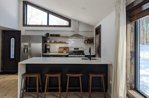 A modern kitchen with a large island, wooden stools, a stainless steel refrigerator, and a window with a view of a snowy landscape. - Acorn Bungalow & Lodge - Luxury Cabin, Catskills (Cairo)