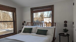 Un dormitorio con una cama grande, dos ventanas, una mesita de noche con lámpara y vistas a árboles y una valla al exterior.