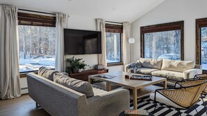 Una sala de estar moderna con un sofá, una mesa de centro y un televisor montado en la pared. Hay ventanas grandes con cortinas, una planta en el alféizar y vistas a un paisaje nevado.
