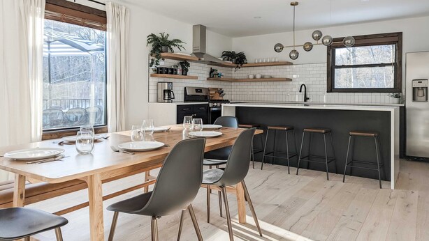A modern kitchen with a wooden dining table, black chairs, and bar stools. There are plants on the shelves, a coffee machine, and a window with a view of trees. - Acorn Bungalow - Luxury Cabin, Catskills (Cairo)