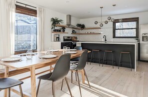 A modern kitchen with a wooden dining table, black chairs, and bar stools. There are plants on the shelves, a coffee machine, and a window with a view of trees. - Acorn Bungalow - Luxury Cabin, Catskills (Cairo)