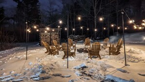 A snow-covered outdoor seating area with wooden chairs and a fire pit, surrounded by string lights. - Acorn Bungalow - Luxury Cabin, Catskills (Cairo)